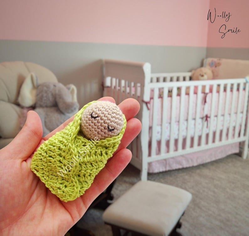 May include: A small crocheted baby doll, wrapped in a green blanket, with closed eyes and a small smile. The doll is being held in a person's hand. The background shows a crib with pink and white bedding.