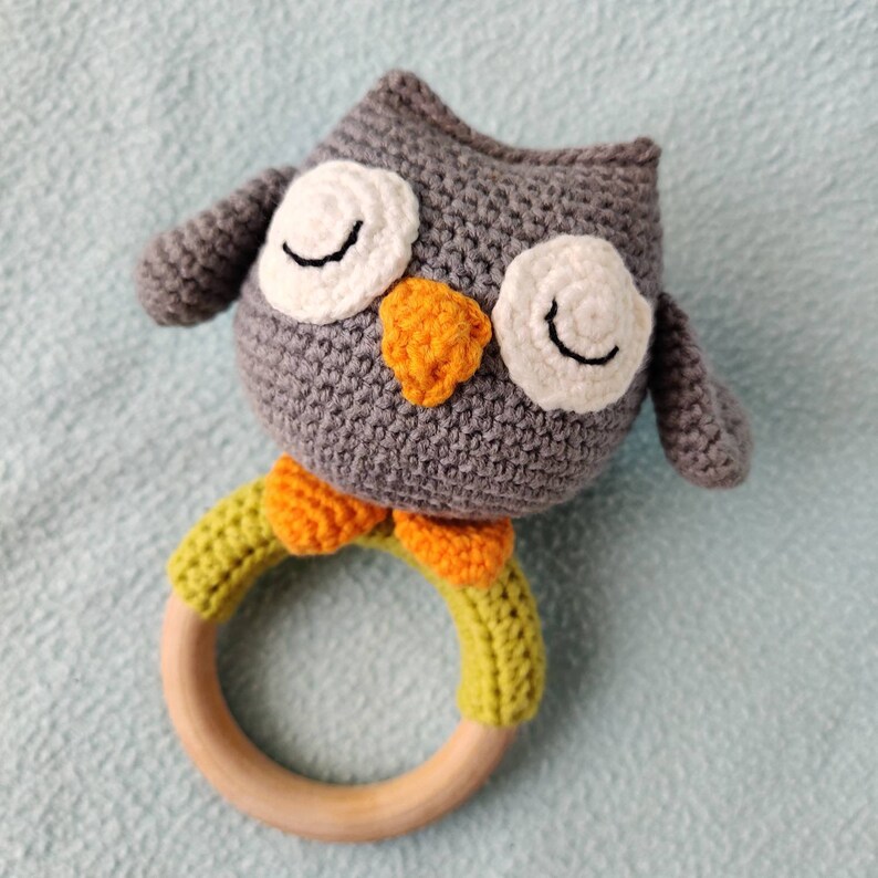 May include: A gray crocheted owl rattle with white eyes and an orange beak. The owl is attached to a wooden ring with green and orange crocheted accents.