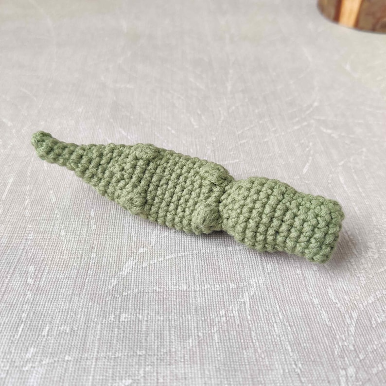 May include: A handmade, light green crochet item, possibly a toy or decorative piece. The item has a textured, bumpy surface and a tapered, pointed end. The crochet work appears tight and even, with a consistent stitch pattern throughout the piece.