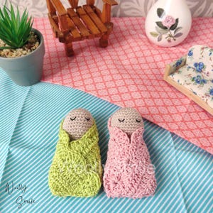 May include: Two crocheted baby dolls, one in green and one in pink, are lying on a blue and white striped blanket. The dolls have closed eyes and are wrapped in blankets.