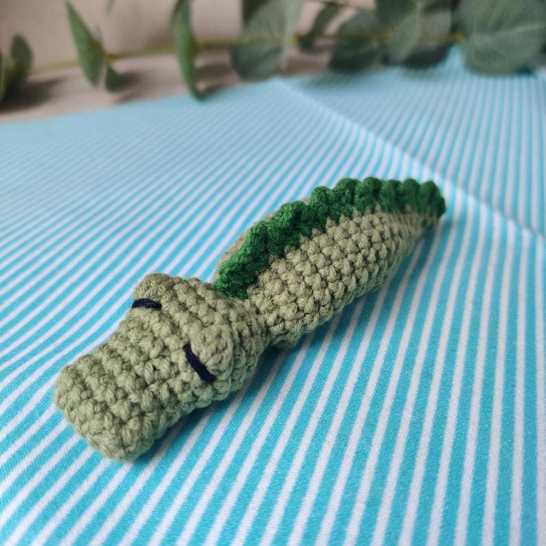 May include: Crocheted alligator toy in shades of green. The alligator has a textured body, a green ridged spine, and small black stitched eyes. The toy is lying on a blue and white striped fabric.