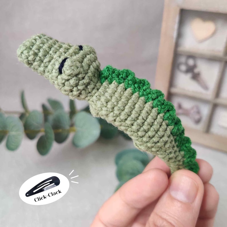 May include: A handmade crochet alligator hair clip in shades of green. The alligator has a detailed textured design, with a darker green spine and a lighter green body. The clip is labeled "Click-Clack".