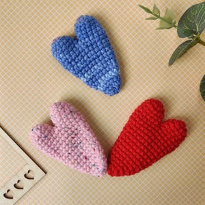 Könnte beinhalten: Drei handgefertigte Häkelherzen in Blau, Rosa und Rot. Das blaue Herz hat ein gesprenkeltes Muster, das rosa Herz hat bunte Flecken und das rote Herz ist einfarbig. Die Herzen sind auf einem gelben Gitterhintergrund angeordnet.