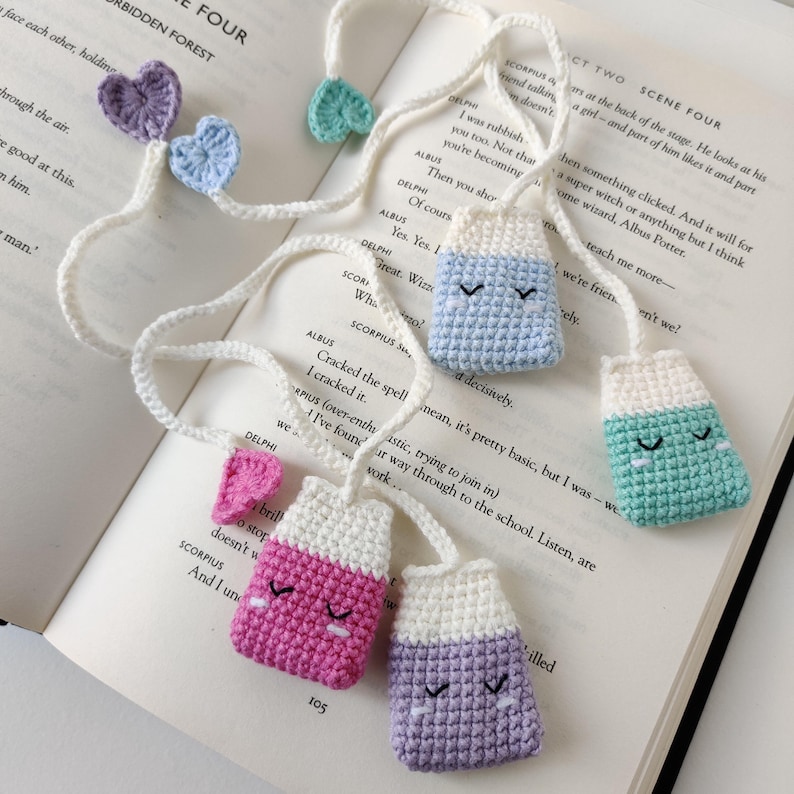 May include: A white string with five crocheted tea bag charms. The charms are in different colours: light blue, light green, pink, purple, and white. Each charm has a white crocheted top and a coloured crocheted bottom. The charms have embroidered eyes and a small white crocheted loop at the top.