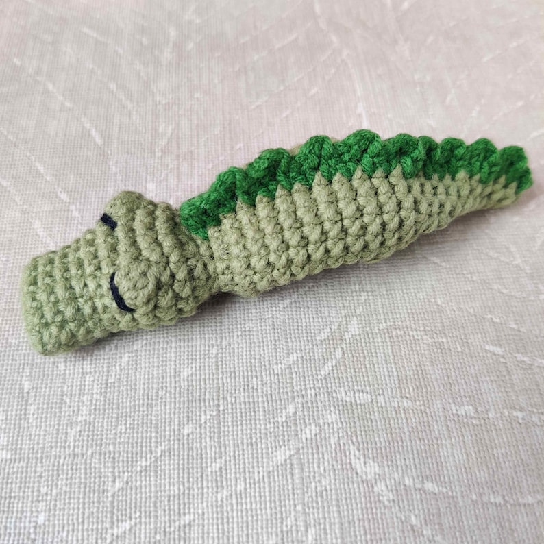 May include: Crocheted alligator toy in shades of green. The alligator has a light green body with a darker green spine and a smiling face. The toy is handmade and appears soft and cuddly.