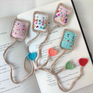 May include: Crocheted bookmarks in the shape of rectangular pastries with colorful sprinkles and smiling faces. Each bookmark is attached to a beige cord, with heart-shaped or leaf-shaped accents. The bookmarks are placed on an open book.