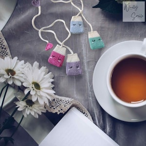 May include: Four crocheted tea bag bookmarks in pink, purple, blue, and green with smiling faces. The bookmarks are hanging from a white string and are on a grey surface with a cup of tea and a book.