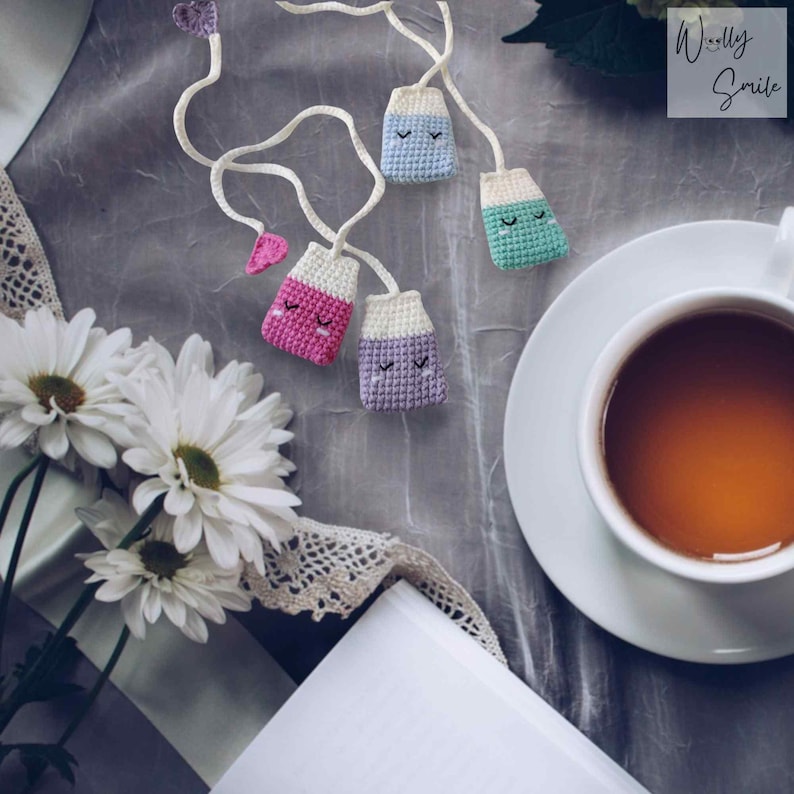 May include: Four crocheted tea bag bookmarks in pink, purple, blue, and green with smiling faces. The bookmarks are hanging from a white string and are on a grey surface with a cup of tea and a book.