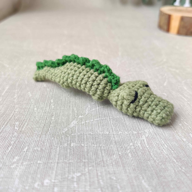 May include: Crocheted alligator toy in shades of green. The body is a light green, with a darker green spine. The alligator has black eyes and is lying on a neutral-colored surface. Handmade crochet animal.
