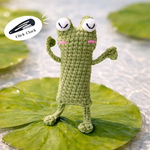 Puede incluir: Una rana de ganchillo verde hecha a mano está sobre una hoja de nenúfar. La rana tiene ojos blancos, mejillas rosadas y brazos extendidos. En la imagen hay una pinza para el pelo negra con el texto "Click-Clack".