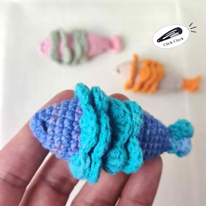 May include: Handmade crochet fish hair clips in blue, pink, and orange. The blue fish clip is in focus, with turquoise fins. The other clips are pink and orange. The text "Click-Clack" is on a black hair clip.