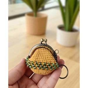 Puede incluir: Un peque&ntilde;o monedero de ganchillo hecho a mano en color amarillo mostaza. El monedero presenta un cierre met&aacute;lico de estilo vintage y est&aacute; adornado con cuentas verdes a lo largo del borde inferior. Se adjunta un llavero de metal.