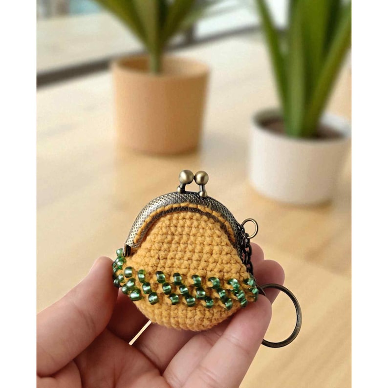 Puede incluir: Un peque&ntilde;o monedero de ganchillo hecho a mano en color amarillo mostaza. El monedero presenta un cierre met&aacute;lico de estilo vintage y est&aacute; adornado con cuentas verdes a lo largo del borde inferior. Se adjunta un llavero de metal.