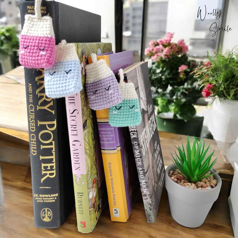 May include: Four crocheted milk carton bookmarks in pink, blue, purple, and green with embroidered eyes and a smiling mouth. The bookmarks are hanging from the spines of four different books.