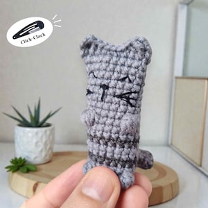 Może przedstawiać: Ręcznie robiony szary szydełkowy kot z czarnymi detalami twarzy, w tym zamkniętymi oczami i wąsami. Kot ma małe, zaokrąglone uszy i łapy. Obraz zawiera tekst "Click-Clack" na czarnej spince do włosów.