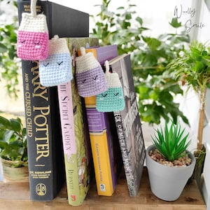 May include: Four crocheted milk carton bookmarks in pink, blue, purple, and green with embroidered eyes and mouths. The bookmarks are hanging from a bookshelf with several books visible. The books include "Harry Potter and the Cursed Child", "The Secret Garden", "Eleanor Hodgman Porter's  The Beck of Mice and Men", and "The Illustrated Classics of  Howells".