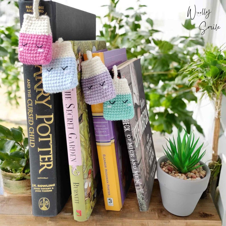 May include: Four crocheted milk carton bookmarks in pink, blue, purple, and green with embroidered eyes and mouths. The bookmarks are hanging from a bookshelf with several books visible. The books include "Harry Potter and the Cursed Child", "The Secret Garden", "Eleanor Hodgman Porter's  The Beck of Mice and Men", and "The Illustrated Classics of  Howells".