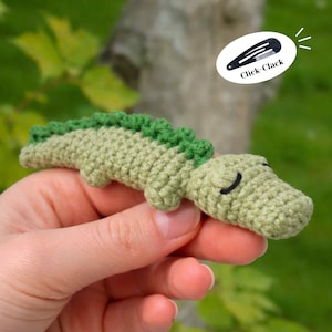 Może przedstawiać: Szydełkowa spinka do włosów w kształcie aligatora w odcieniach zieleni. Aligator ma teksturowane ciało, ciemniejszy zielony grzbiet i zamknięte oczy. Klips jest oznaczony "Click-Clack".