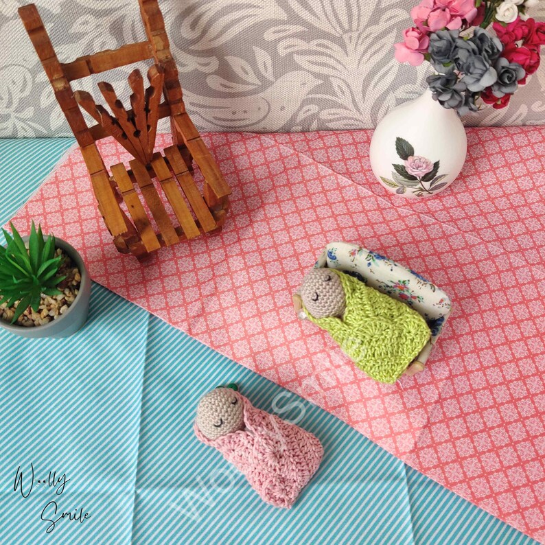 May include: Two crocheted baby dolls, one in a pink swaddle and one in a green swaddle, are lying on a pink and white patterned surface. The doll in the green swaddle is lying in a miniature floral patterned bed.