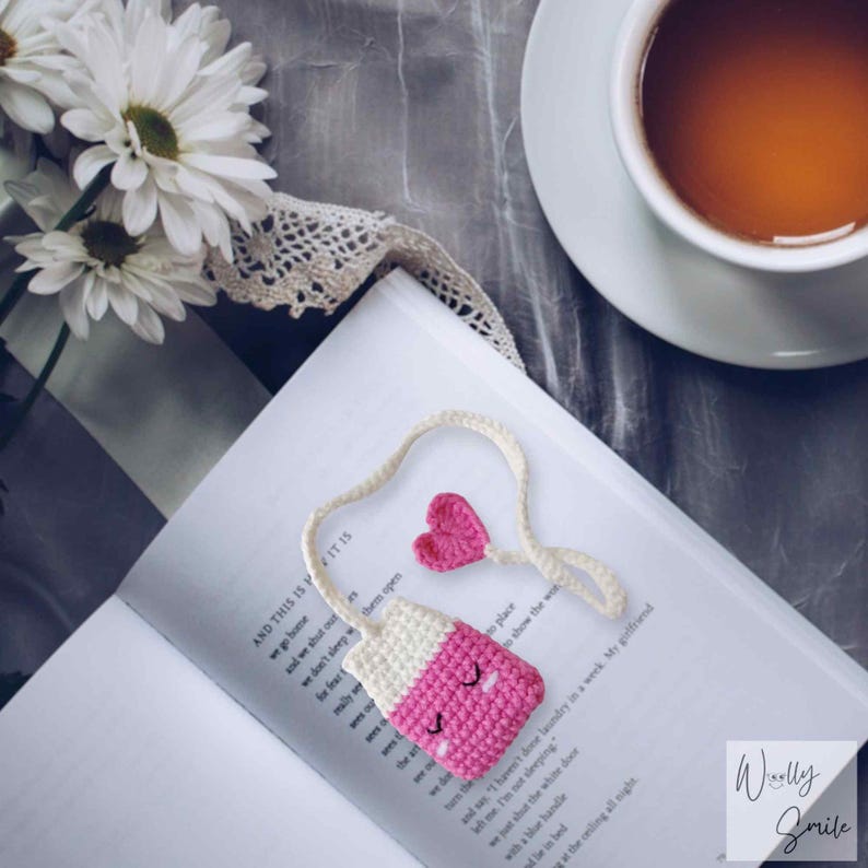 May include: A pink and white crocheted tea bag charm with a heart-shaped charm attached to a white cord. The charm is sitting on a book with the text "AND THIS IS IT IS" visible. The text "Wolly Smile" is visible in the bottom right corner of the image.