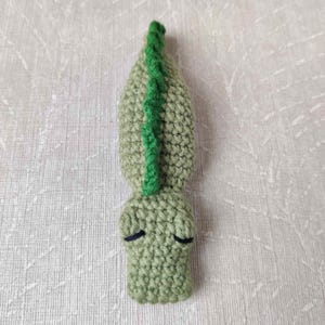 May include: A handmade, light green crocheted alligator with a bright green spine. The alligator has closed eyes and is made of soft yarn. The item is a unique, handcrafted piece.
