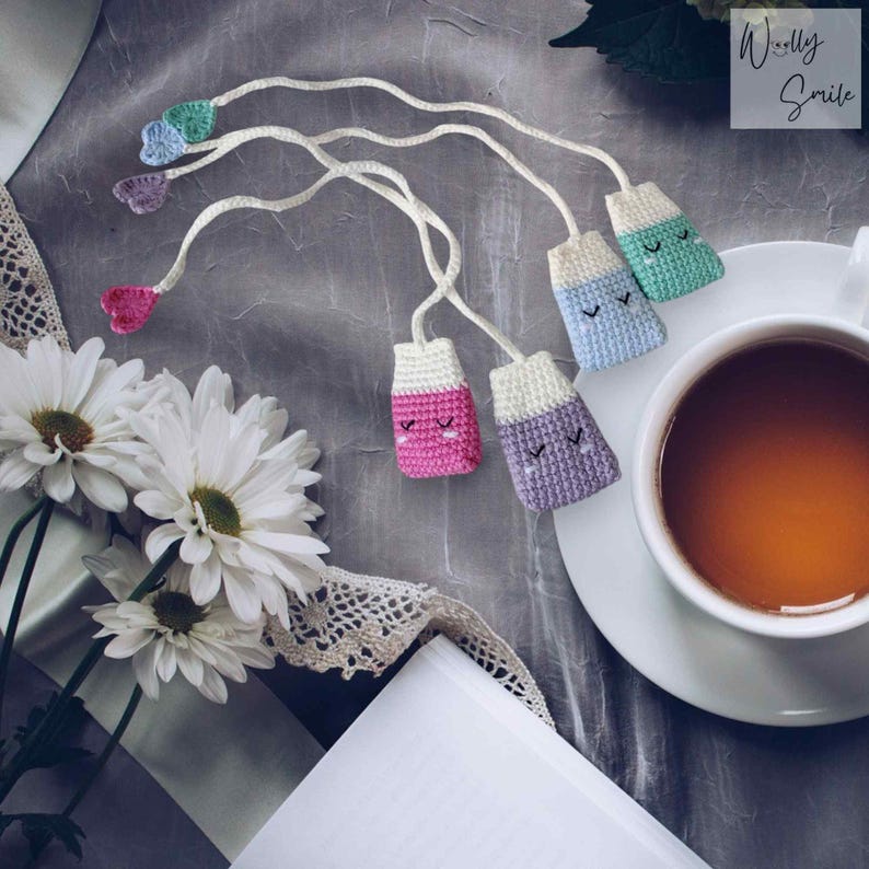 May include: Four crocheted tea bag bookmarks with smiling faces. The bookmarks are in pink, purple, blue, and green. The bookmarks are hanging from a white string and are on a grey background. The text "Woolly Smile" is in the top right corner of the image.