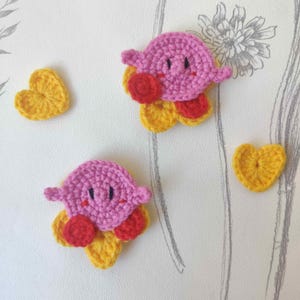 May include: Crocheted pink character with red accents and yellow flower-like base. Two yellow crocheted hearts are also present. The items are handmade and likely part of a craft project or decorative set.