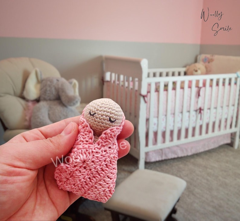 May include: A small crocheted baby doll in a pink swaddle, with closed eyes, lying in a hand. The doll is in front of a white crib with pink and white bedding.