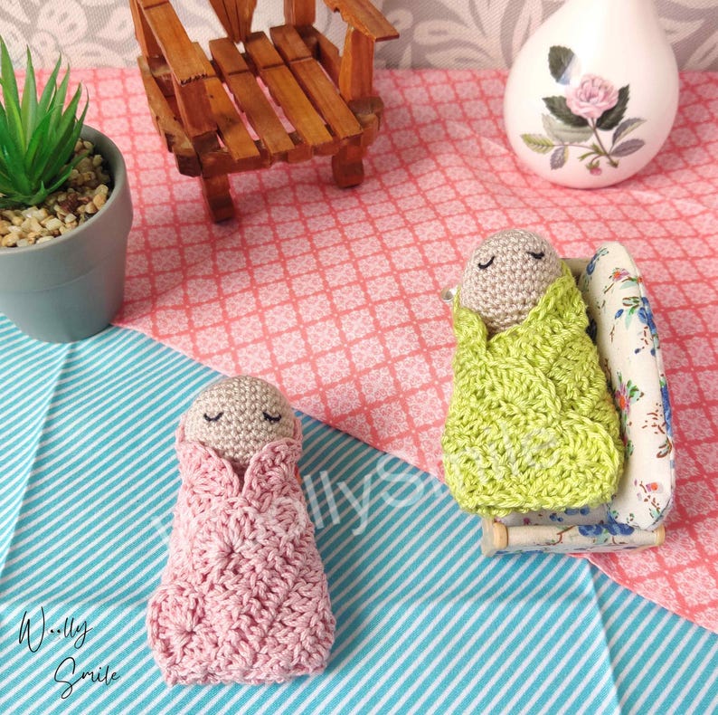 May include: Two crocheted baby dolls, one in a pink blanket and one in a green blanket, are lying on a striped blue and white surface. The doll in the green blanket is lying on a miniature floral couch.