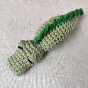 May include: A handmade crochet alligator in shades of green. The body is a light green, with a darker green spine detail. The alligator has a small head with black stitched eyes and a smiling mouth. The toy is made of soft yarn.