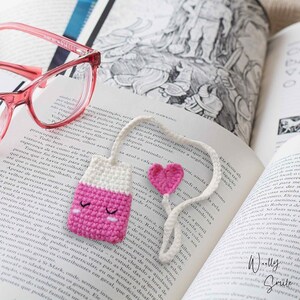 May include: A pink and white crocheted bookmark shaped like a small bottle with a pink heart attached to the top. The bookmark is lying on an open book.