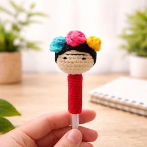 May include: A handmade crochet pen featuring a doll's head with a black hair and colorful flower crown. The pen's body is red, and the doll's face is beige. The pen is held in a hand.