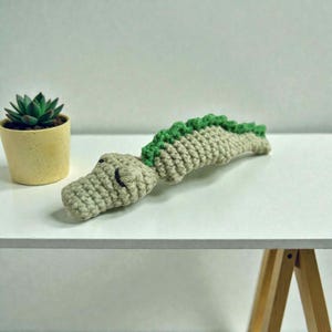 May include: A crocheted alligator toy in shades of beige and green. The alligator has a textured body with a green spine and a detailed head. The toy is lying on a white surface, with a small potted succulent plant in the background.