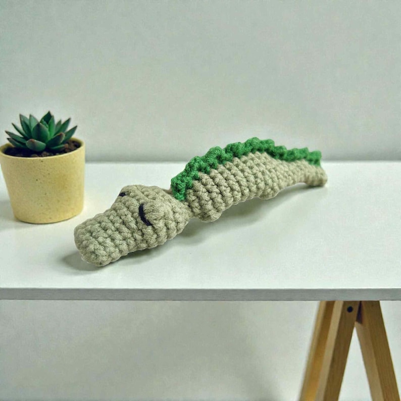May include: A crocheted alligator toy in shades of beige and green. The alligator has a textured body with a green spine and a detailed head. The toy is lying on a white surface, with a small potted succulent plant in the background.