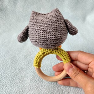 May include: A gray crocheted owl rattle with a wooden ring and green crocheted accents. The owl is attached to the ring.
