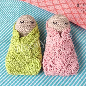May include: Two crocheted baby dolls, one in pink and one in green, with closed eyes. The dolls are wrapped in crocheted blankets.
