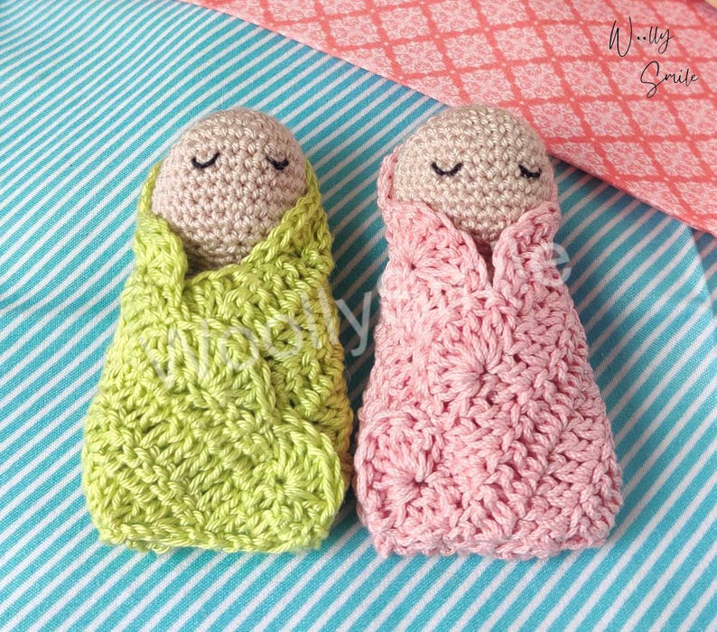 May include: Two crocheted baby dolls, one in pink and one in green, with closed eyes. The dolls are wrapped in crocheted blankets.