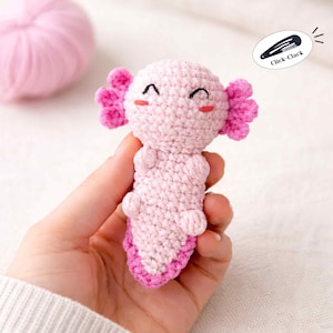 May include: A handmade, light pink crochet axolotl with bright pink gills and accents. The axolotl has a smiling face with black stitched eyes and rosy cheeks. A ball of pink yarn is in the background. The image also includes a black hair clip with the text "Click-Clack".
