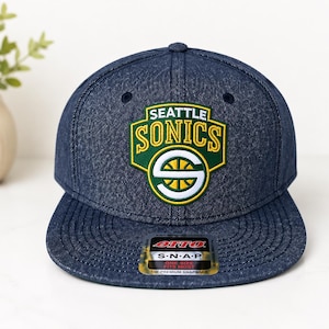 May include: A denim blue snapback hat featuring the Seattle Sonics logo in green, yellow, and white. The logo is a shield shape with the team name and a basketball design. The hat has a flat brim and an "OTTO" tag.