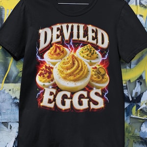 May include: Black t-shirt featuring a graphic of deviled eggs with the text "DEVILED EGGS" in a bold, retro font. The design includes images of deviled eggs with various toppings and lightning bolt accents.