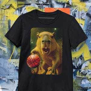 May include: Black t-shirt featuring a capybara holding a basketball. The capybara has an open mouth and is set against a background with a basketball court. The shirt is made of a soft material.