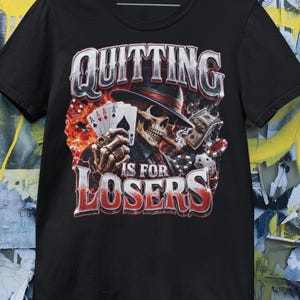 May include: A black short-sleeve t-shirt featuring a graphic print of a skeleton wearing a hat and smoking a cigar, holding playing cards. The text reads "QUITTING IS FOR LOSERS" in bold, distressed lettering, surrounded by dice and poker chips.