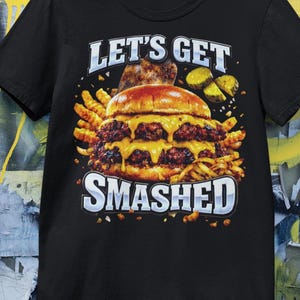 Puede incluir: Camiseta negra con un gráfico de una hamburguesa doble con queso derretido, patatas fritas y pepinillos. El texto "LET'S GET SMASHED" se muestra arriba y abajo de la hamburguesa en blanco.