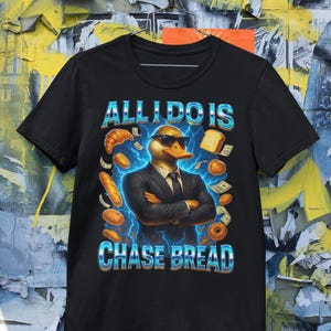 May include: Black t-shirt featuring a duck wearing sunglasses and a suit, with the text "ALL I DO IS CHASE BREAD". The duck is surrounded by images of bread, money, and lightning bolts.