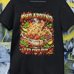 Peut inclure: T-shirt noir avec un graphisme coloré d'un bol de pho, de baguettes et de flammes. Le texte "PHO AROUND" est au-dessus du bol et "AND FIND OUT" en dessous. Le design comprend de la vapeur, du basilic et du citron vert.