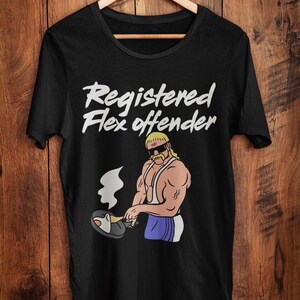 May include: Black t-shirt with the text "Registered Flex Offender" in white script. The shirt features a cartoon illustration of a muscular man with a blonde mullet, sunglasses, and a mustache, cooking in a pan.
