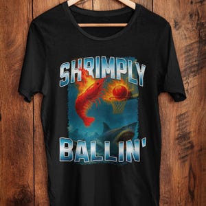 May include: Black t-shirt with a humorous graphic. The design features a red shrimp dunking a basketball into a hoop, with a shark below. The text reads "SHRIMPLY BALLIN'" in a blue and white outlined font.