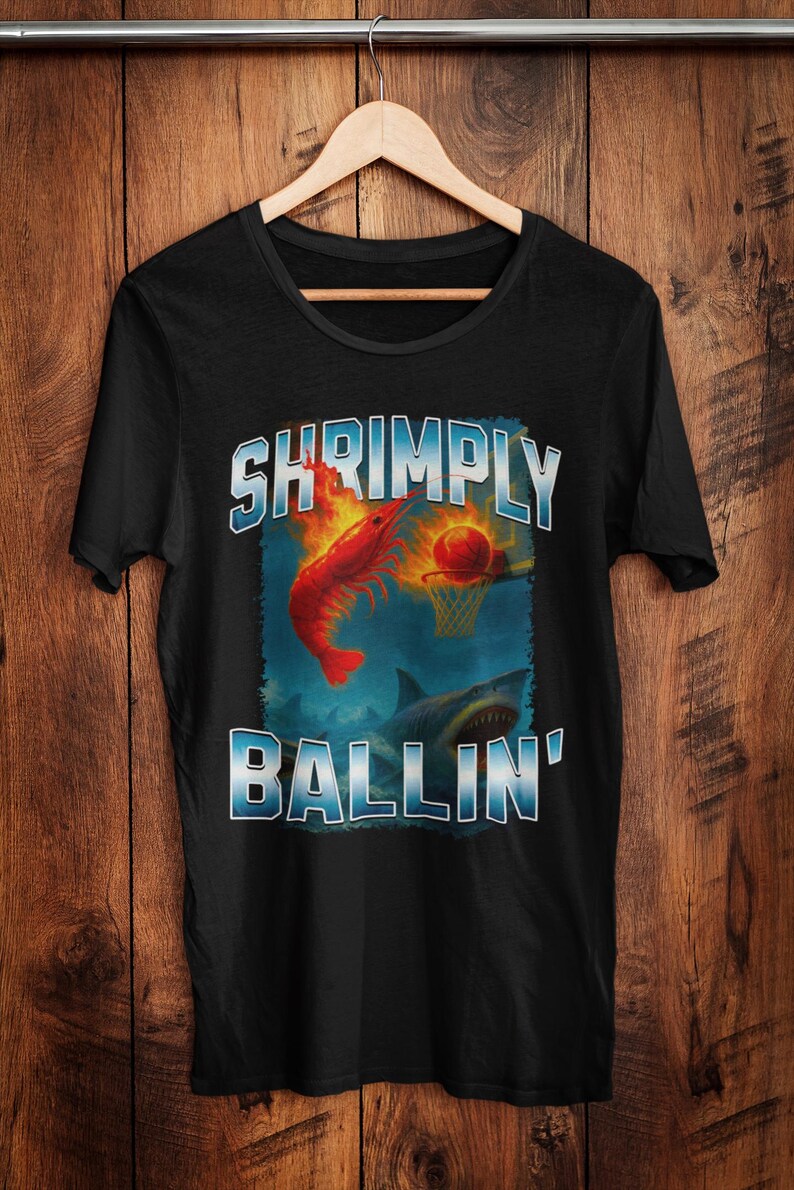 May include: Black t-shirt with a humorous graphic. The design features a red shrimp dunking a basketball into a hoop, with a shark below. The text reads "SHRIMPLY BALLIN'" in a blue and white outlined font.