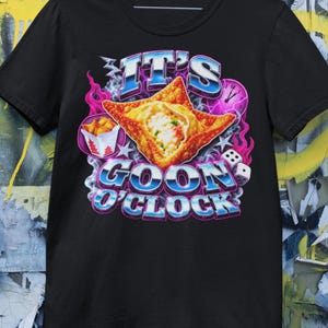 Puede incluir: Una camiseta negra con un estampado gráfico de un wonton frito, una caja de comida para llevar, dados y un reloj. El texto IT'S GOON O'CLOCK aparece en una fuente azul metálica. El diseño incluye llamas rosas, humo y estrellas.