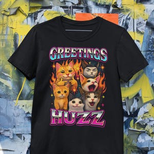 Op de afbeelding: Zwart t-shirt met een kleurrijke afbeelding van katten met vlammen en de tekst "GREETINGS HUZZ". Het ontwerp heeft een retro-stijl en de katten hebben verschillende uitdrukkingen.
